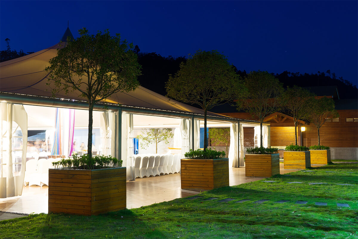 Vue nocturne d’une tente éclairée avec des arbres en pot alignés devant, dans un cadre extérieur.