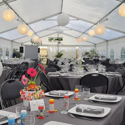 L'intérieur d'une tente de réception avec des tables rondes couvertes de nappes grises. Les tables sont décorées avec des assiettes, des verres, des couverts et des centres de table colorés. Des lanternes blanches sont suspendues au plafond, créant une am