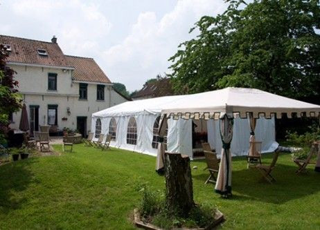 Un grand chapiteau blanc installé sur une pelouse verte, entouré d'arbres et d'arbustes. Le chapiteau est ouvert sur un côté, révélant l'intérieur avec des tables et des chaises, prêt pour un événement en plein air.
