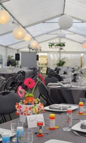 L'intérieur d'une tente décorée pour un événement festif. Les tables sont couvertes de nappes grises et sont décorées avec des serviettes et des sets de table à motifs tropicaux. Au centre des tables, il y a des arrangements floraux avec des fleurs exotiq