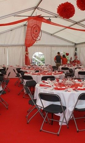 L'intérieur d'une tente décorée pour un événement. Les tables sont couvertes de nappes blanches et sont entourées de chaises noires. Les tables sont décorées avec des arrangements floraux et des décorations rouges suspendues au plafond. La tente est insta
