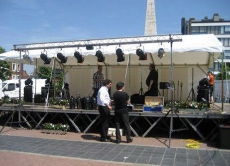 Une scène de concert en plein air avec un chapiteau blanc en arrière-plan. La scène est équipée de plusieurs projecteurs et d'équipements audio. Des musiciens sont sur scène, prêts à jouer devant un public.
