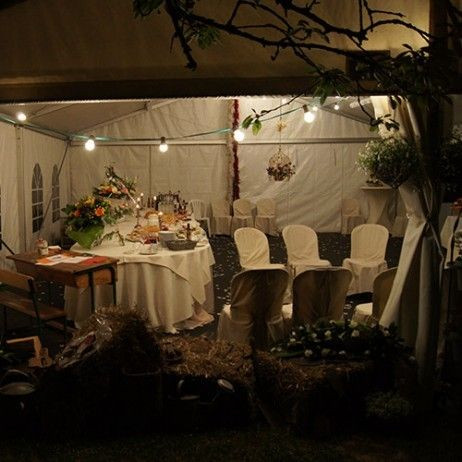 L'intérieur d'une tente décorée pour un événement festif. Les tables sont couvertes de nappes blanches et sont entourées de chaises avec des housses blanches. Les tables sont décorées avec des arrangements floraux et des bougies, créant une ambiance roman