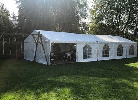 Un grand chapiteau blanc installé dans un jardin bien entretenu. Le chapiteau est entouré de plusieurs arbres et arbustes, et il y a des fenêtres en forme d'arcs sur le côté. À proximité, on peut voir une maison et un abri de jardin en bois. La pelouse es