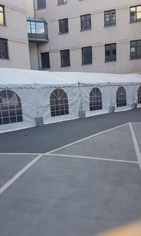 Un grand chapiteau blanc installé sur une surface pavée dans un environnement urbain, entouré de bâtiments. Le chapiteau a des fenêtres en forme d'arcs sur le côté, laissant entrer la lumière naturelle.