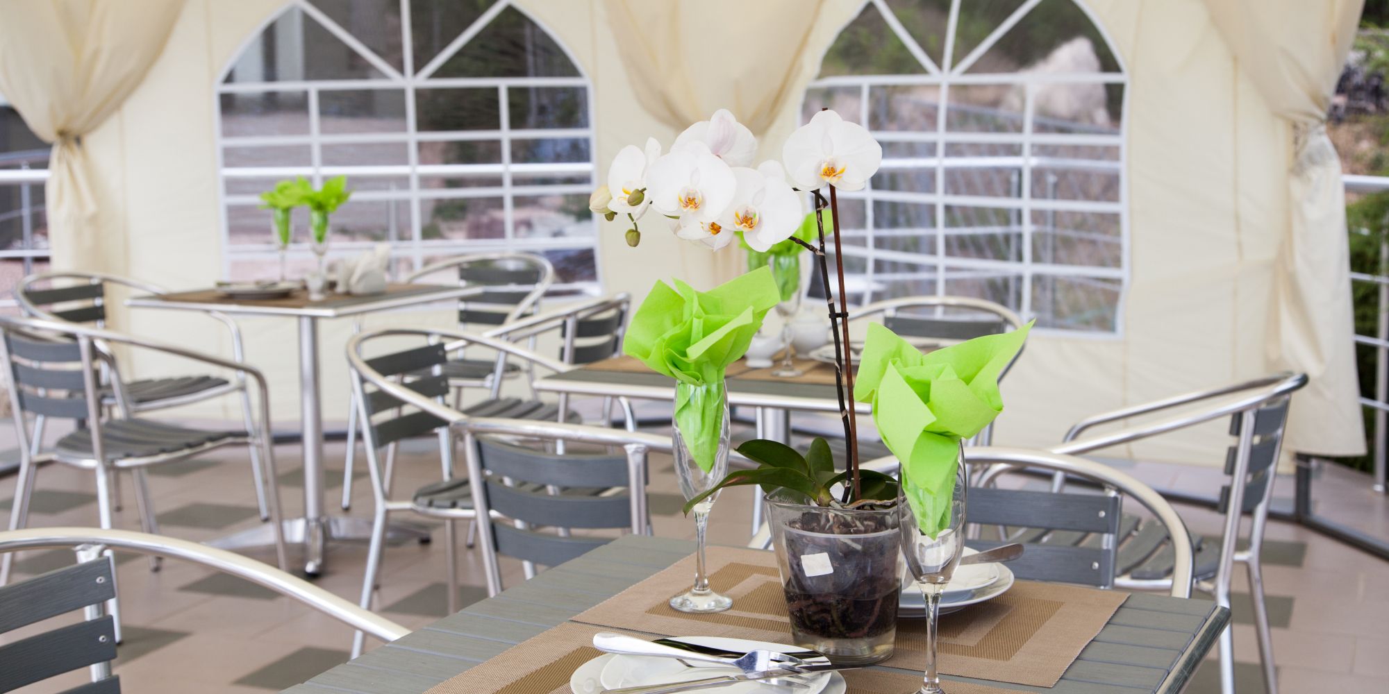 Une table dans un espace extérieur couvert, décorée avec une plante en pot et des serviettes vertes. La table est entourée de chaises métalliques et est préparée pour un repas.