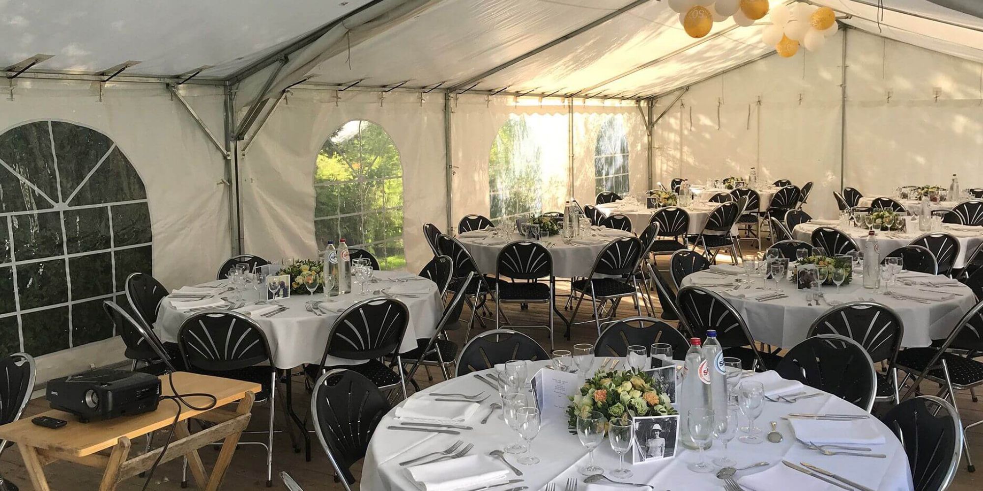 Une salle de réception dans une tente avec des tables rondes couvertes de nappes blanches et des chaises noires. Des ballons dorés sont suspendus au plafond, et un projecteur est installé pour une présentation.