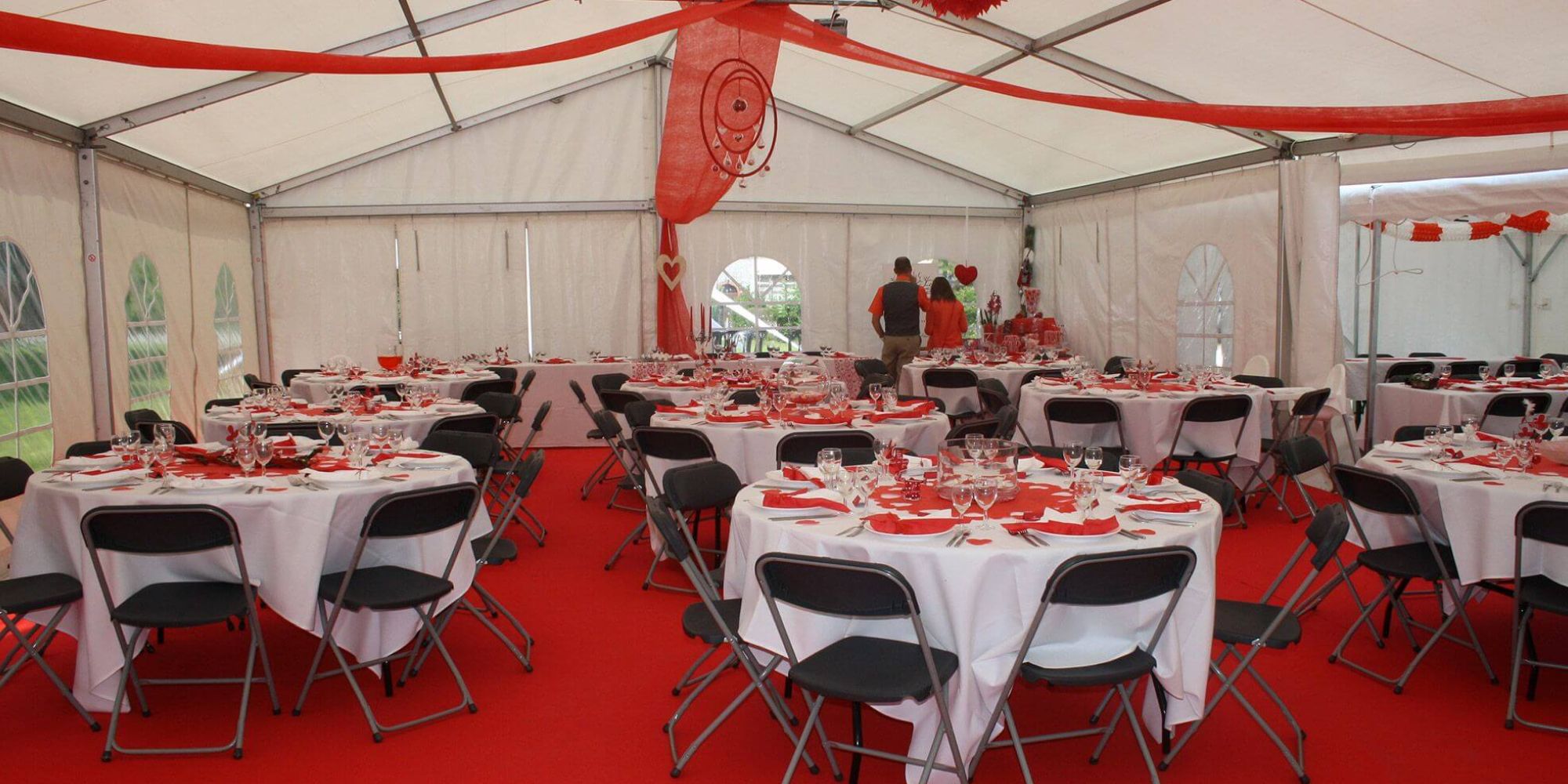 L'intérieur d'une grande tente de réception décorée avec des nappes rouges et des chaises noires. Des guirlandes rouges pendent du plafond, et des tables sont disposées pour un événement festif.
