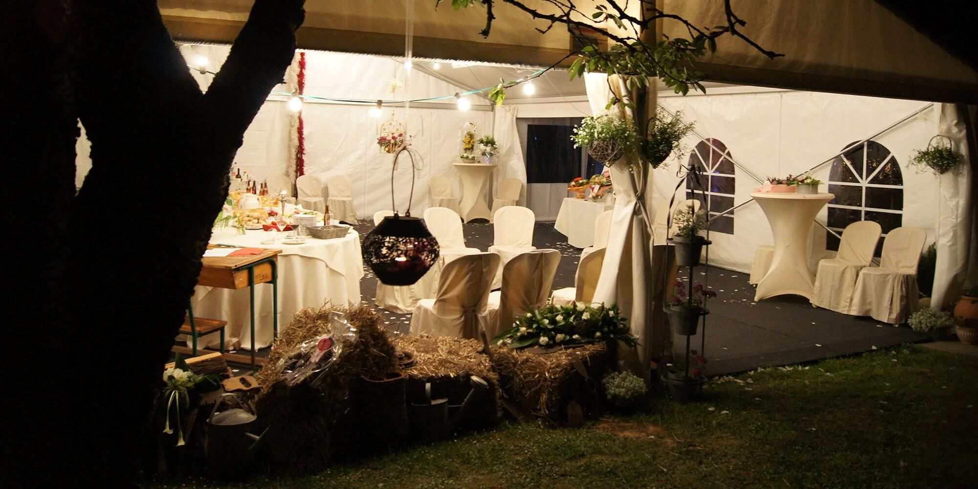 Une vue nocturne d'une tente de réception avec des tables couvertes de nappes blanches et des chaises assorties. Des décorations suspendues et des plantes ajoutent une touche festive à l'ambiance.