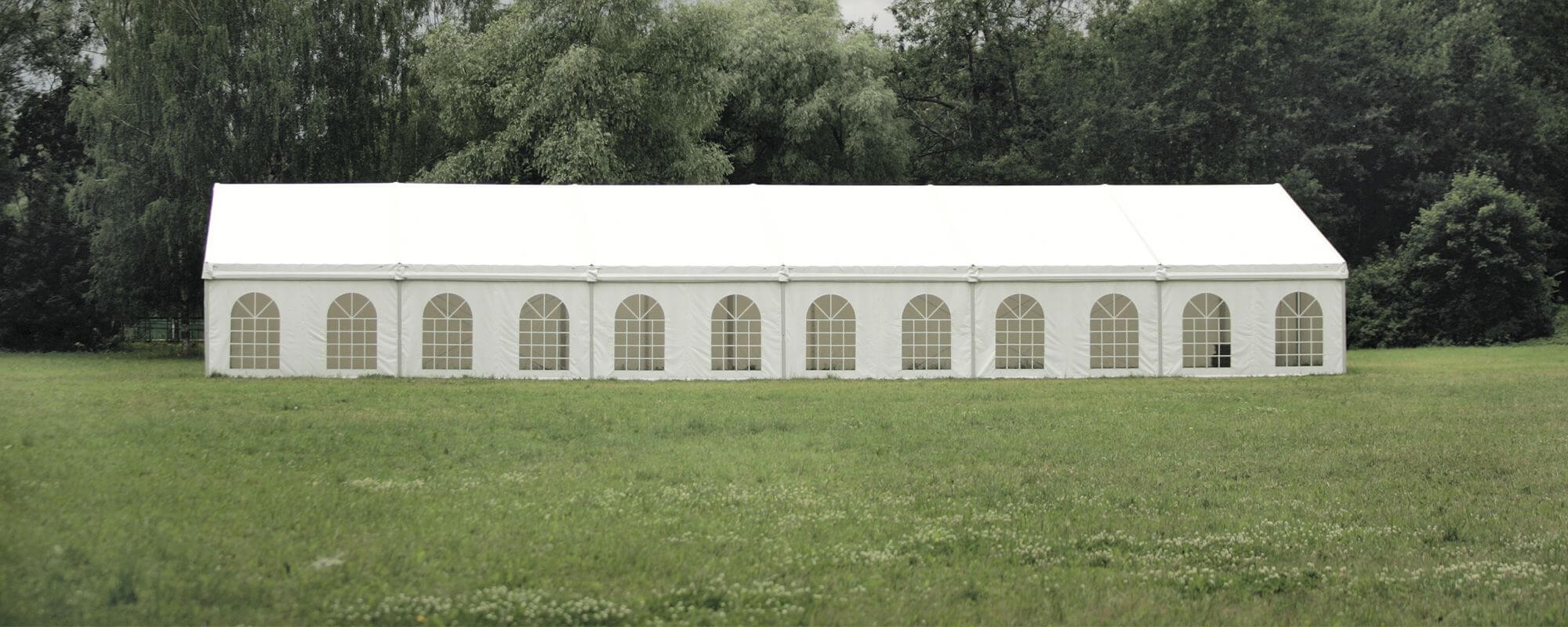 Un grand chapiteau blanc installé sur un champ herbeux, entouré d'arbres et de verdure. Le chapiteau a plusieurs fenêtres en forme d'arcs sur le côté, laissant entrer la lumière naturelle.