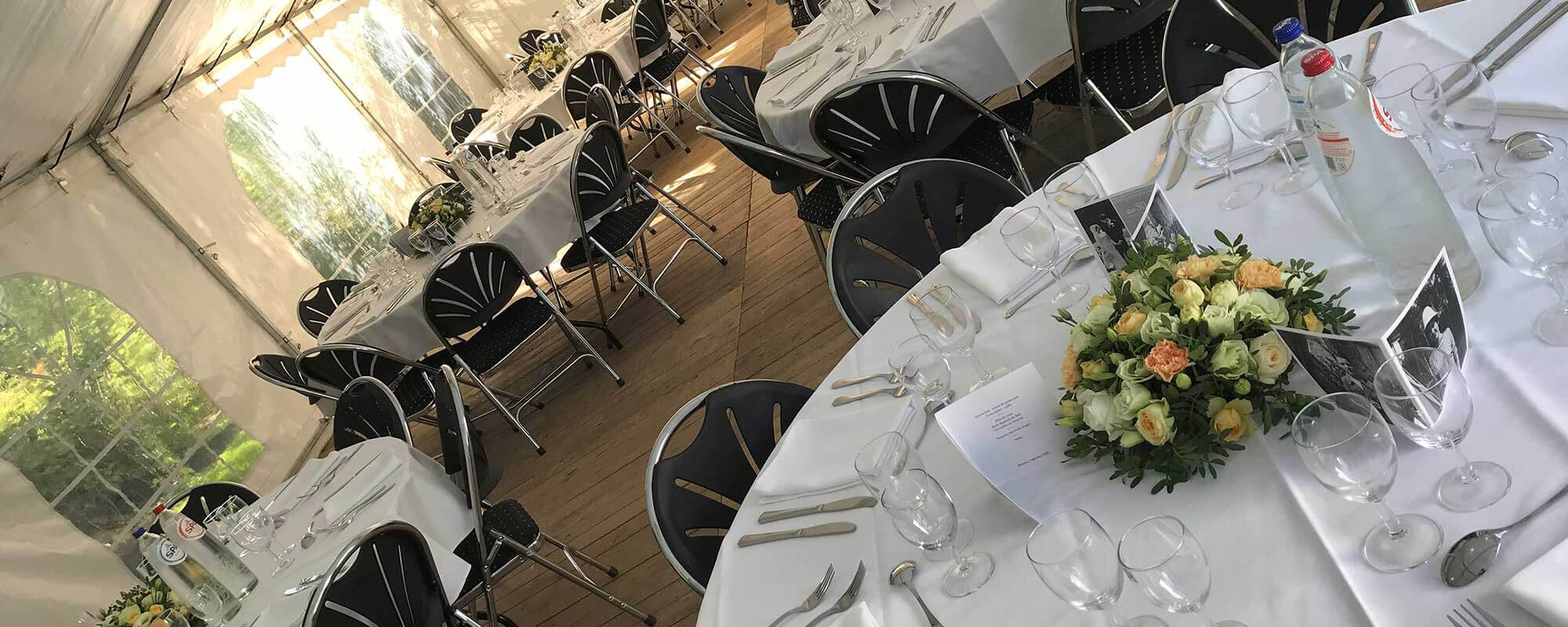 L'intérieur d'une tente de réception avec plusieurs tables rondes couvertes de nappes blanches. Chaque table est décorée avec des bouquets de fleurs et des cadres photo, et est entourée de chaises noires. Les tables sont également équipées de verres, d'as