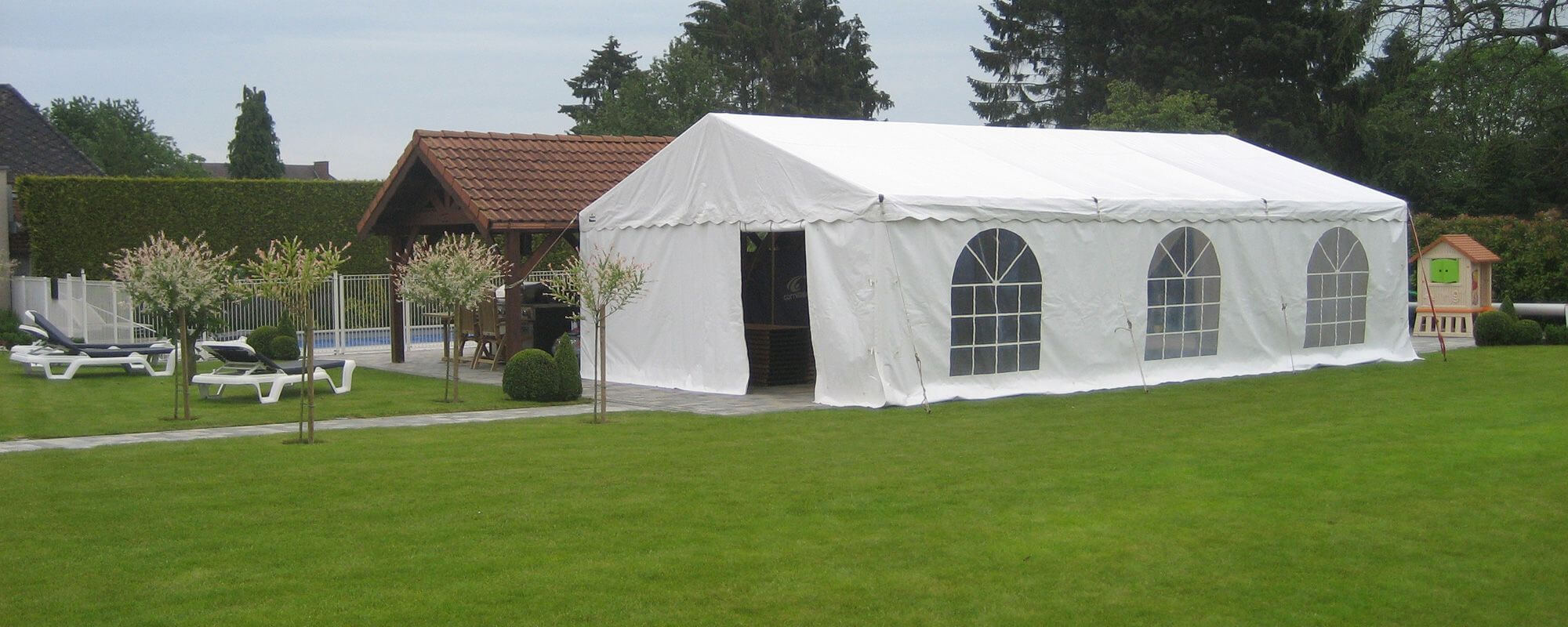 Un grand chapiteau blanc installé dans un jardin bien entretenu. Le chapiteau est entouré de plusieurs arbres et arbustes, et il y a des fenêtres en forme d'arcs sur le côté. À proximité, on peut voir une piscine avec des chaises longues et un abri de jar